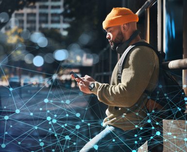 A man outside in a city typing into his cellphone. A network of blue lines suggesting a network is coming out of the phone and spreading across the image.