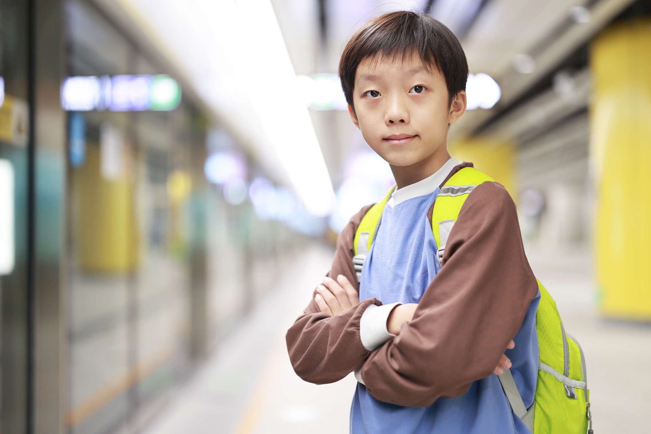 A schoolaged boy wearing a yellow backpack waits on a train platform with his arms crossed in front of him.