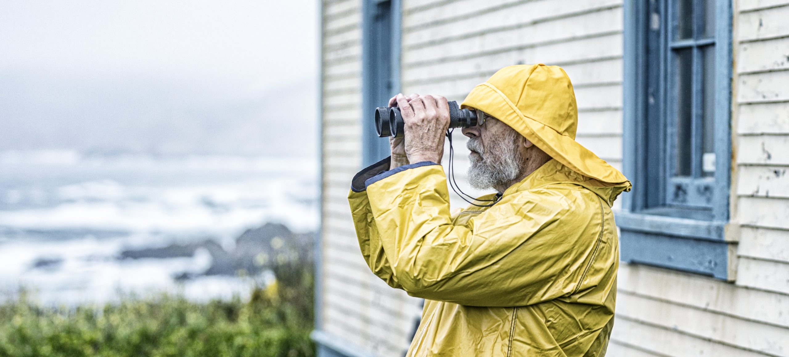 A senior adult ancient mariner sailor gray beard lighthouse keeper man is looking through binoculars out over the Pacific Ocean surf. He is standing next to his lighthouse residence building located on the California coastline. He is wearing a bright yellow waterproof fisherman's rain hat and matching rain slicker raincoat rain jacket. 