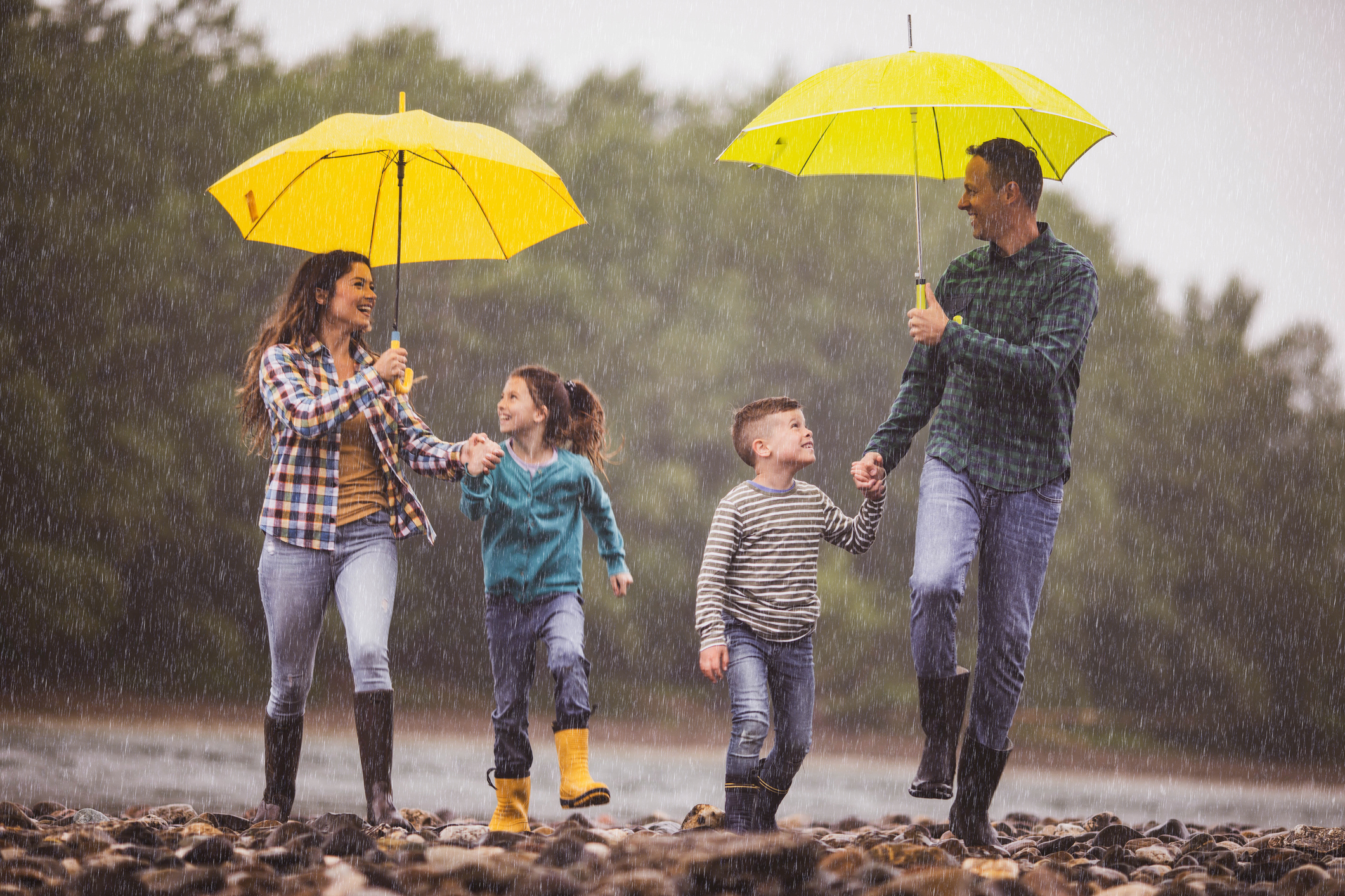 Carefree parents and their kids having fun while holding hands and running on a rainy day by the river. Parents are holding umbrellas.