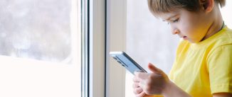 Little toddler boy is holding the smartphone at home. He is wearing bright yellow t-shirt. Playing video games like a social problem