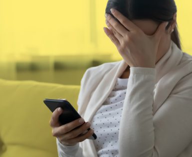 Woman sitting on a couch holding a cellphone and covering her face with one hand