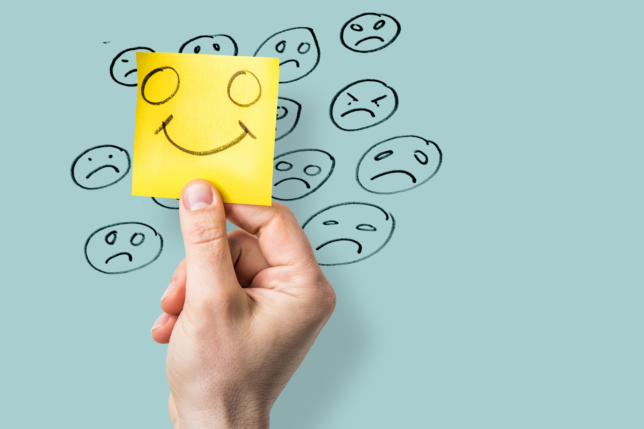 hand holding yellow notepaper showing a happy face over unhappy  emoticons drawn on blue background