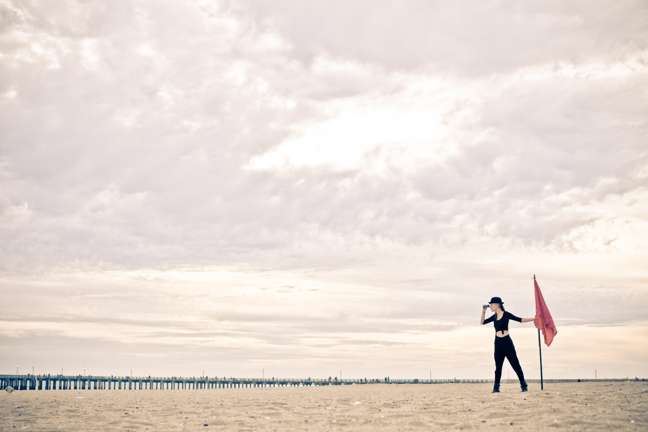 A woman dressed all in black stands with her binoculars next to a giant red flag she placed in the sand of her newly discovered land (aka Coney Island)