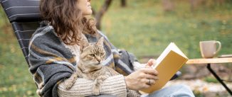 Young woman sitting in the autumn garden with a pet gray cat in her hands reading literature