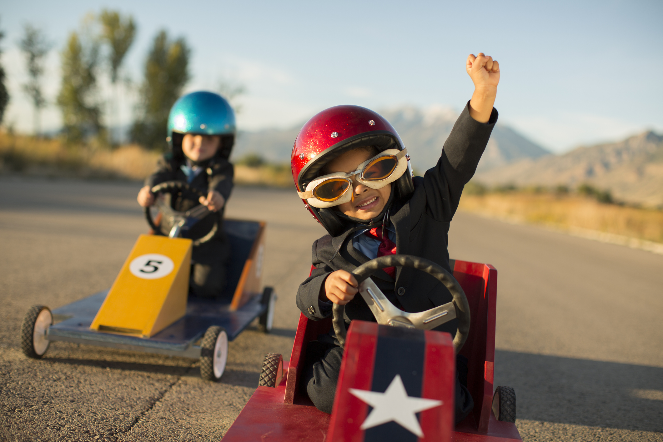 two young children racing in pretend racecars. The winner is raising a fist in the air.