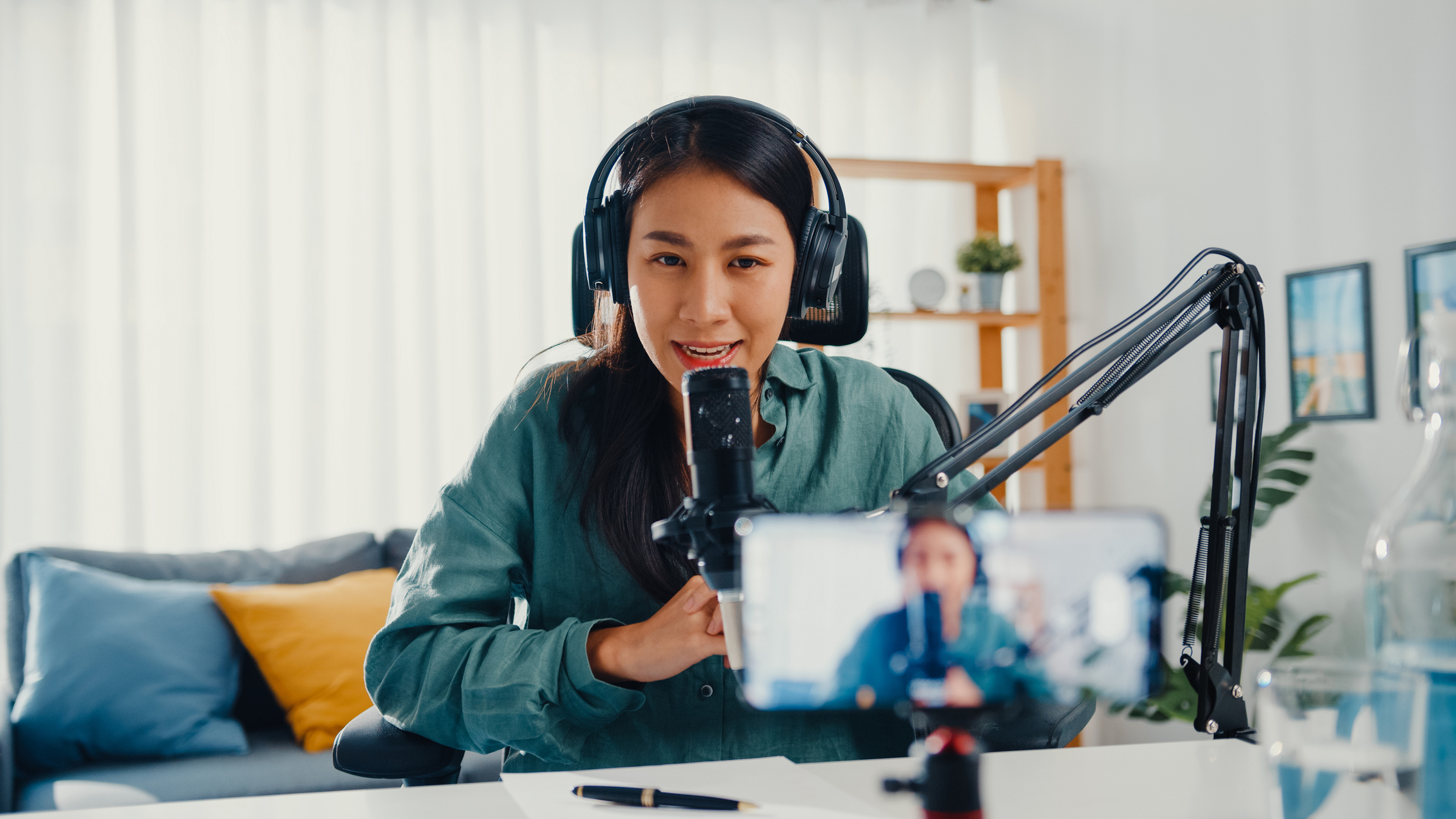 Teenage Asia girl influencer use microphone wear headphone record content with smart phone for online audience listen at house. Student female podcaster make audio podcast from her home studio.