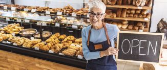 Portrait of a happy senior business owner holding an open sign at a cafe and smiling - food and drinks concepts.