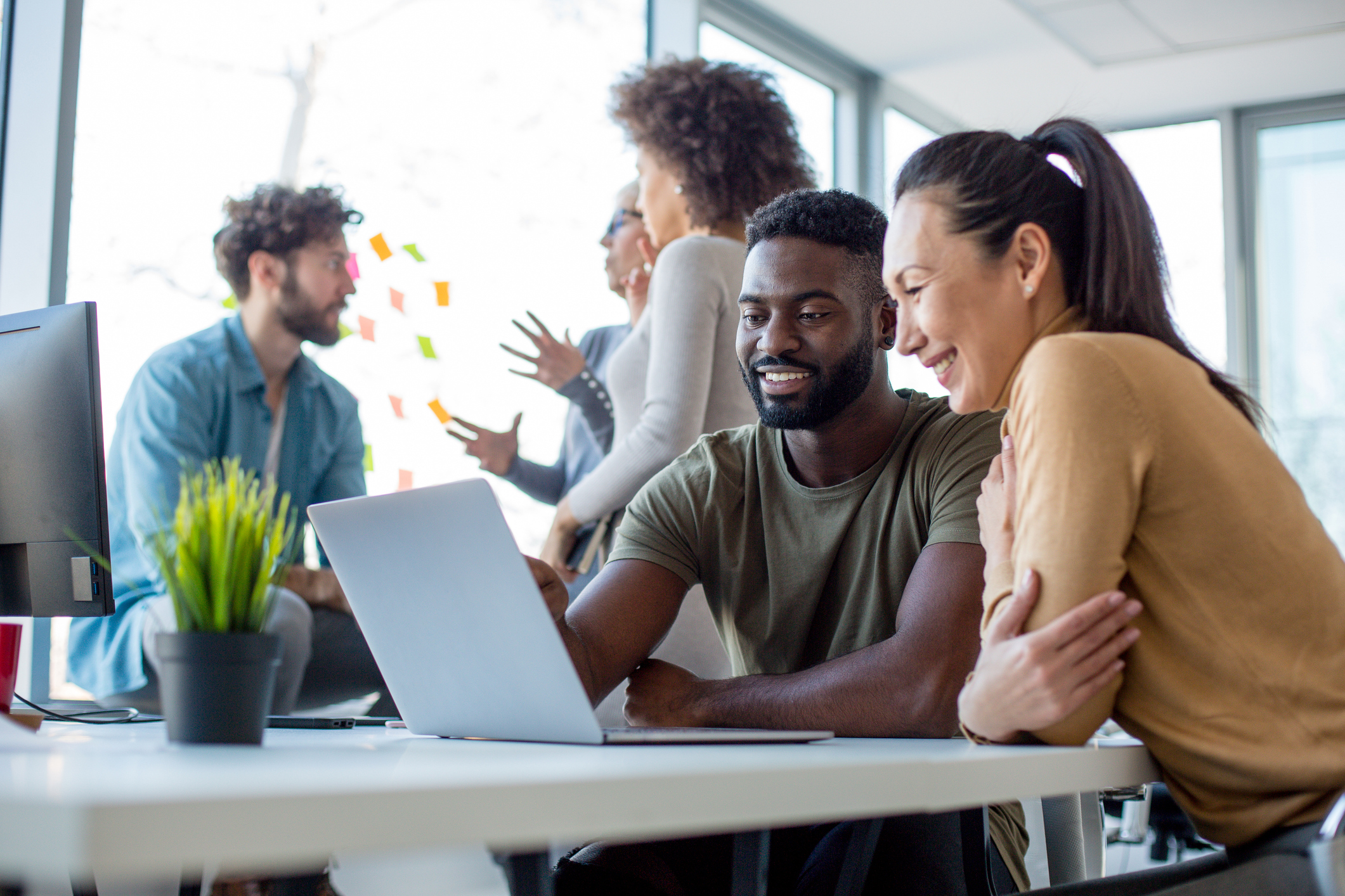 A man and woman in an office setting look at a laptop screen and smile at what they see.