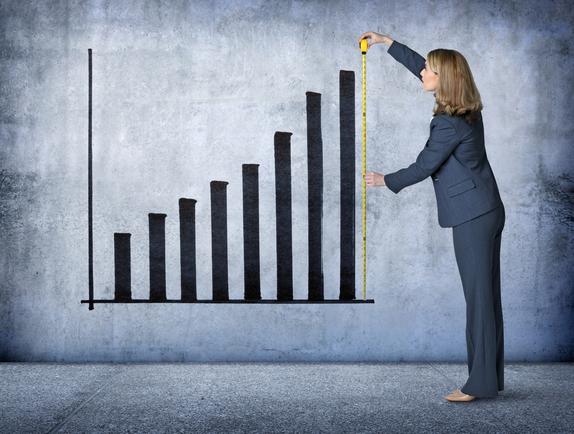 A businesswoman uses a tape measure to measure the growth of an upward pointing graph projected on a concrete wall.