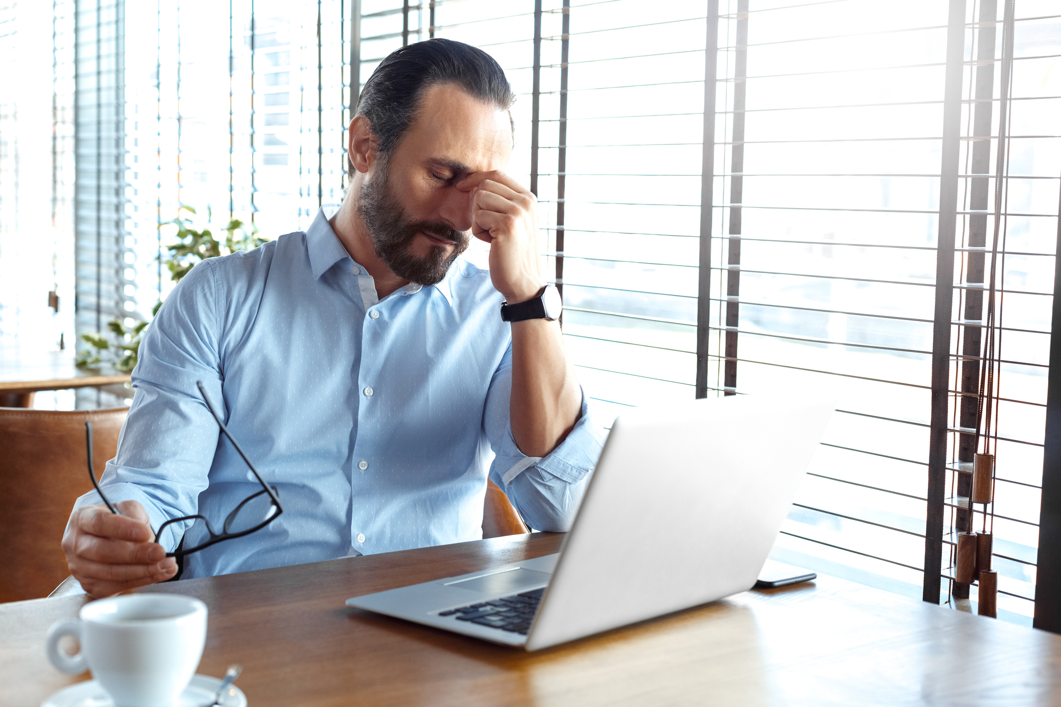 Mature man trader sitting at table at cafe at daytime with cup of coffee and laptop holding eyeglasses closed eyes massaging nose bridge tired