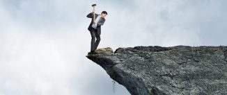 A businessman swings a sledge hammer against a rocky cliff as he stands on the wrong side placing himself at great risk.