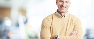 A mature businessman wearing casual clothes while standing arms crossed in the office.