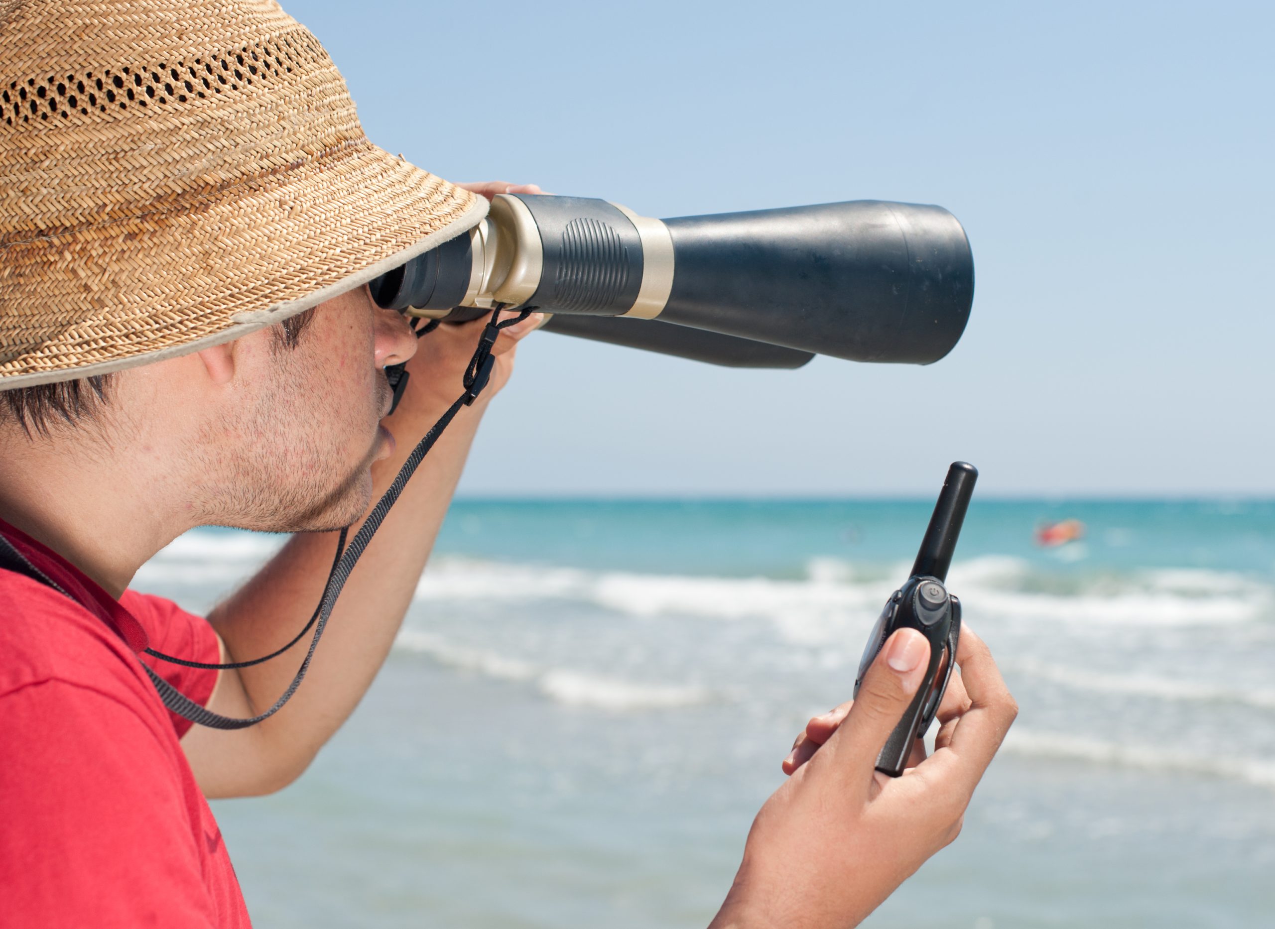 A man looking at the ocean through binoculars while holding a walkie-talkie.