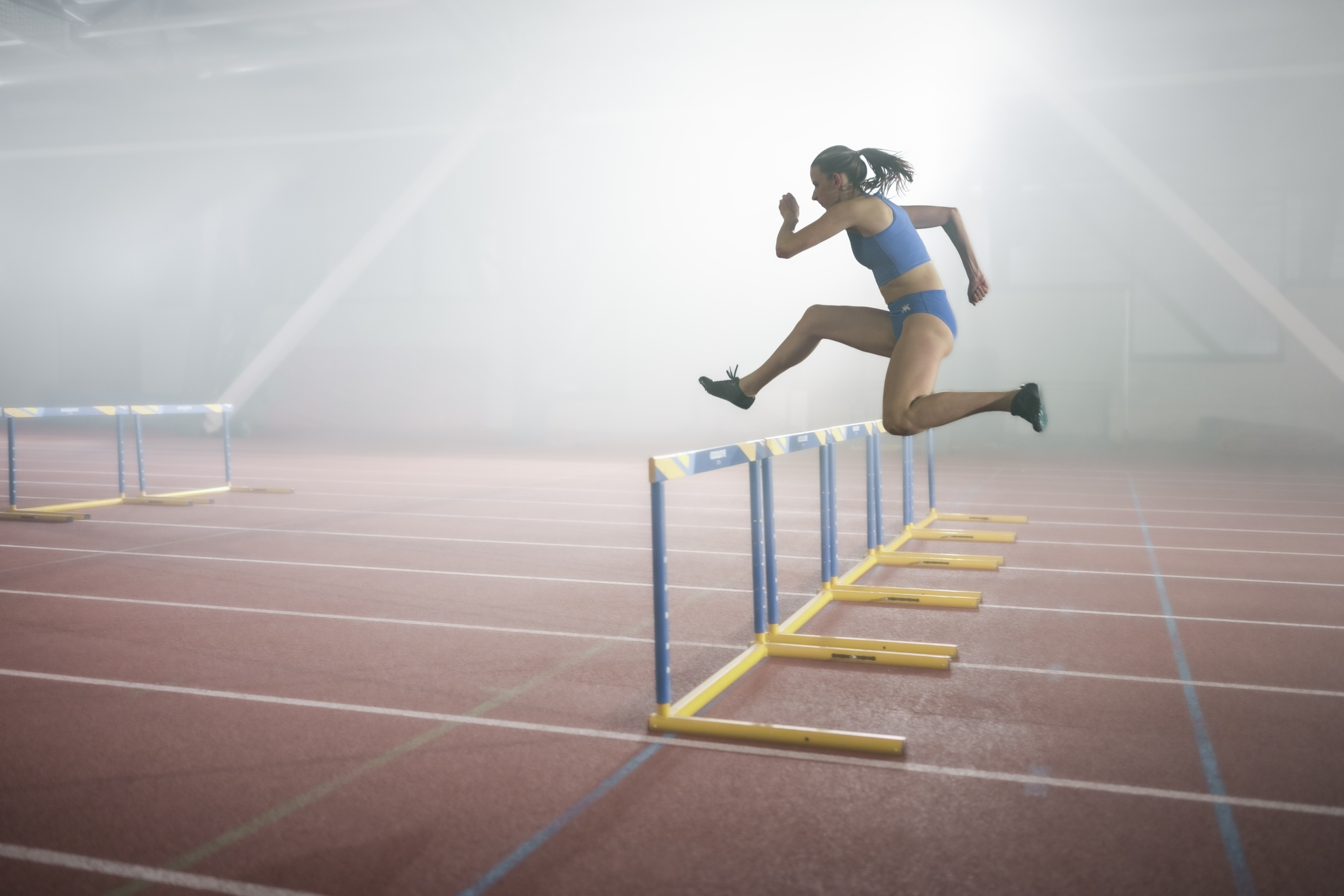 Side view of athlete young woman jumping over hurdles during practice in sports hall.