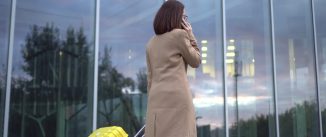 A young woman walks with a yellow suitcase and speaks on the phone. A girl with glasses and a coat walks along the mirrored building. 4k