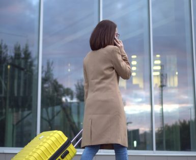 A young woman walks with a yellow suitcase and speaks on the phone. A girl with glasses and a coat walks along the mirrored building. 4k