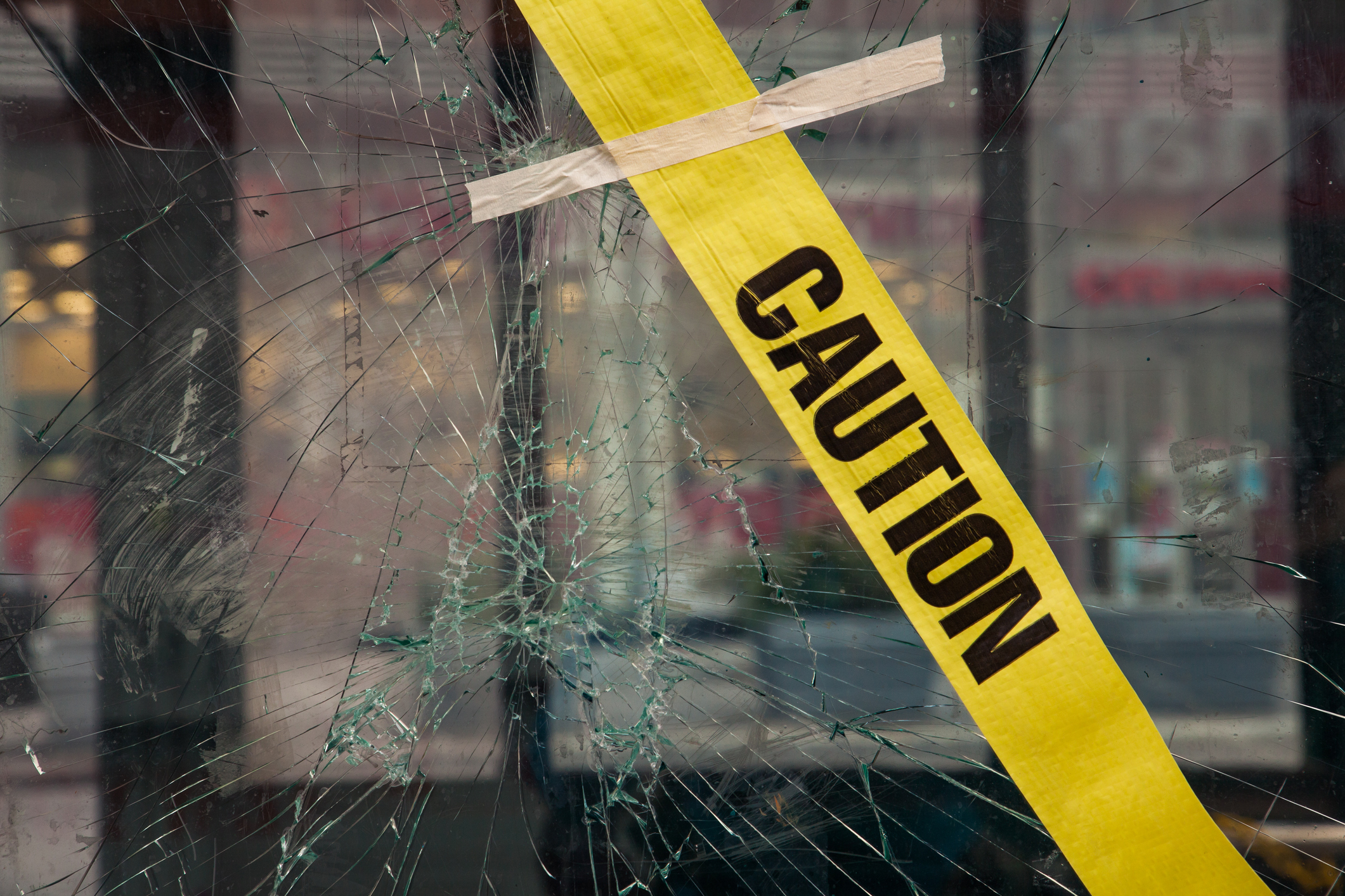 Cracks radiate from the center of the glass where the impact was made. Down town shop windows in the background in strong blur. Yellow warning tape with the text "CAUTION" is taped to the glass. Rainy day. NYC. Downtown Manhattan. New York. USA